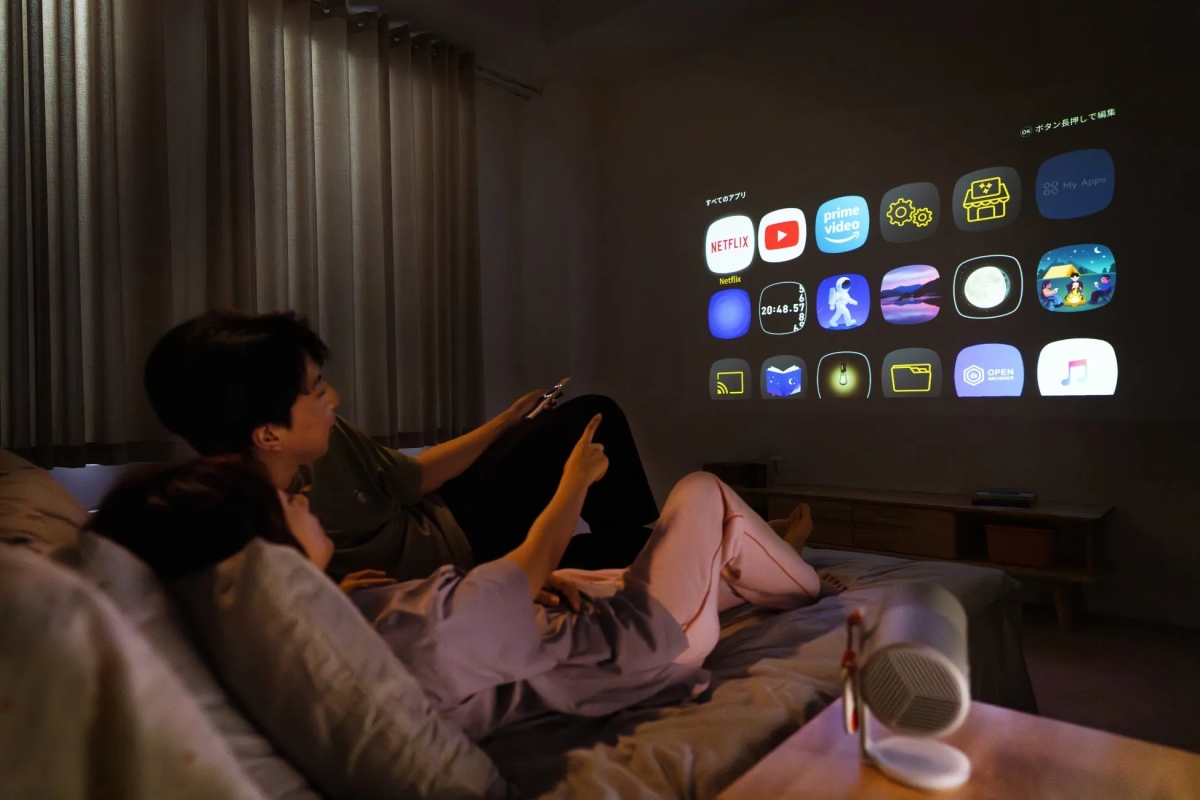 ベッドでくつろぎながらプロジェクターで映像を見ている2人。壁にはアプリのアイコンが表示されており、NetflixやYouTubeなどのサービスが利用できることがわかる。