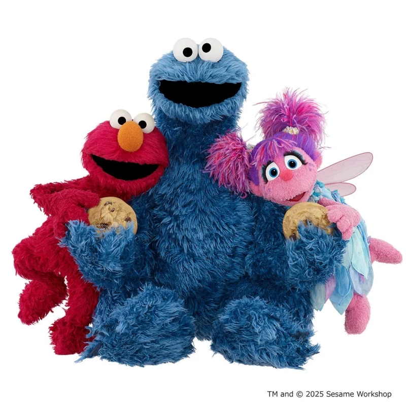 セサミストリートのキャラクター、エルモ、クッキーモンスター、アビー・カダビーが寄り添っている画像です。エルモとアビーはクッキーを手に持っています。