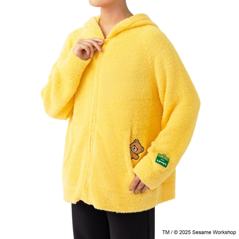 黄色いフード付きのセサミストリートのキャラクターがデザインされたパーカーを着ている人の写真です。パーカーにはポケットがあり、クマのキャラクターのパッチが付いています。