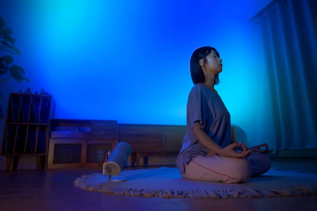 女性が瞑想している写真。青い光に照らされた部屋で、床に座り、目を閉じています。部屋には、植物、収納棚、加湿器のようなものが写っています。