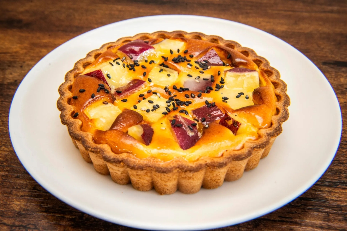 お皿に載った、さつまいもとパイナップルのタルトのクローズアップ写真。タルトは、さつまいもとパイナップルの塊、黒いゴマがトッピングされています。背景は木製の表面です。