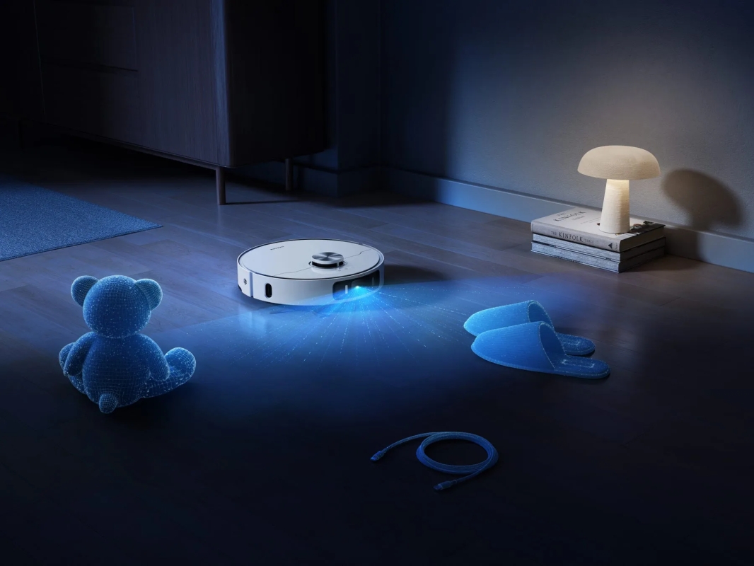 夜の部屋で、ロボット掃除機が床を掃除している様子。床には、クマのぬいぐるみ、スリッパ、ケーブル、本が置かれている。
