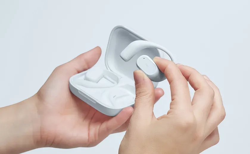 白い充電ケースから、JBLのロゴが入ったライトグレーのオープンイヤー型ワイヤレスイヤホンを取り出している様子。