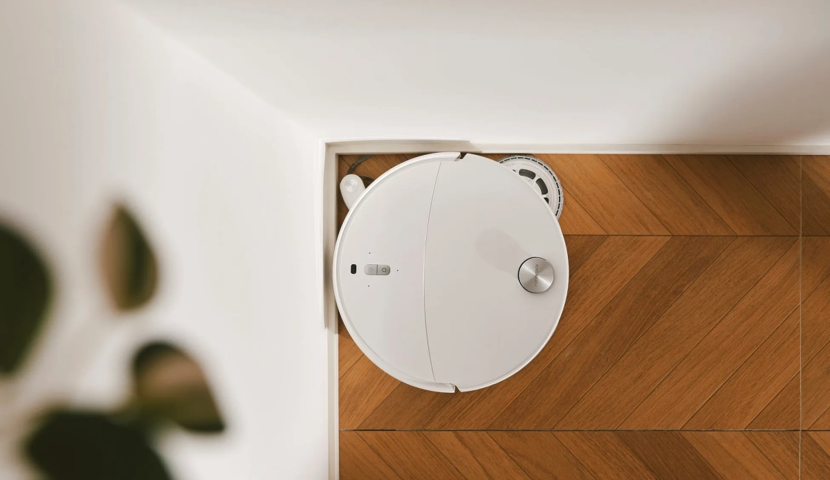 白いロボット掃除機が木製の床に置かれています。上部からのショットで、背景に白い壁があります。