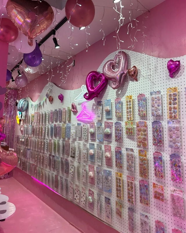 ピンク色の内装の店内で、壁には様々なステッカーが陳列されており、上部にはハート型の風船や照明が飾られています。