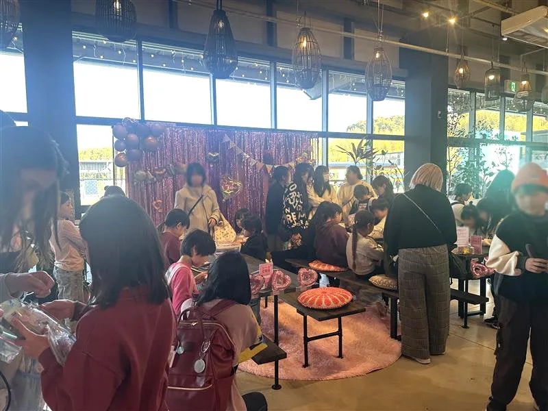 カフェのような場所で、子供たちが集まってイベントを行っている様子。ピンク色の装飾やバルーンがあり、誕生日会のような雰囲気も感じられる。