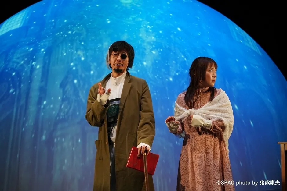 舞台に立つ2人の俳優。男性は赤い本と杖を持ち、女性は白いショールを羽織っている。背景は青色の星空のようなデザイン。