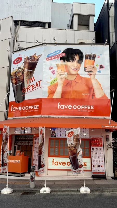 カフェ「fave COFFEE」の屋外広告と店舗の写真。広告には、JAY CHANGがコーヒーカップを持ち笑顔で写っており、「Save COFFEE with JAY CHANG」という文字とコーヒーの写真が描かれている。店舗はオレンジ色を基調とし、コーヒーのメニューやロゴが掲示されている。