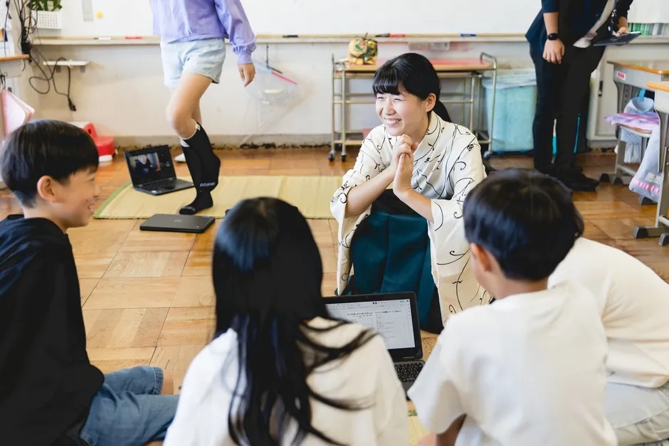 教室で、笑顔の女性と子供たちがノートパソコンを見ながら話している様子。