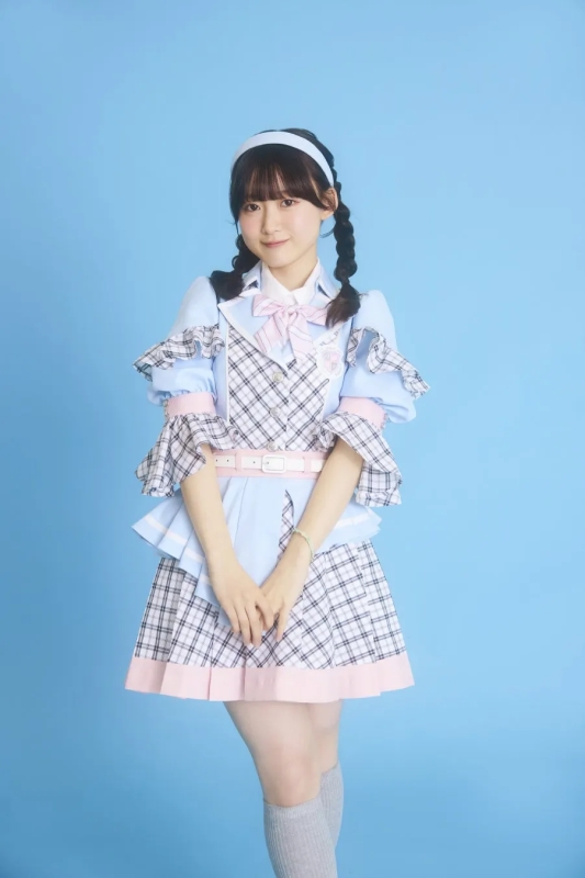 水色の衣装を着た女性のポートレート。彼女は、チェック柄のミニスカート、ピンクのリボン、髪に白いヘッドバンド、そして三つ編みをしている。彼女はカメラを見て微笑んでいる。