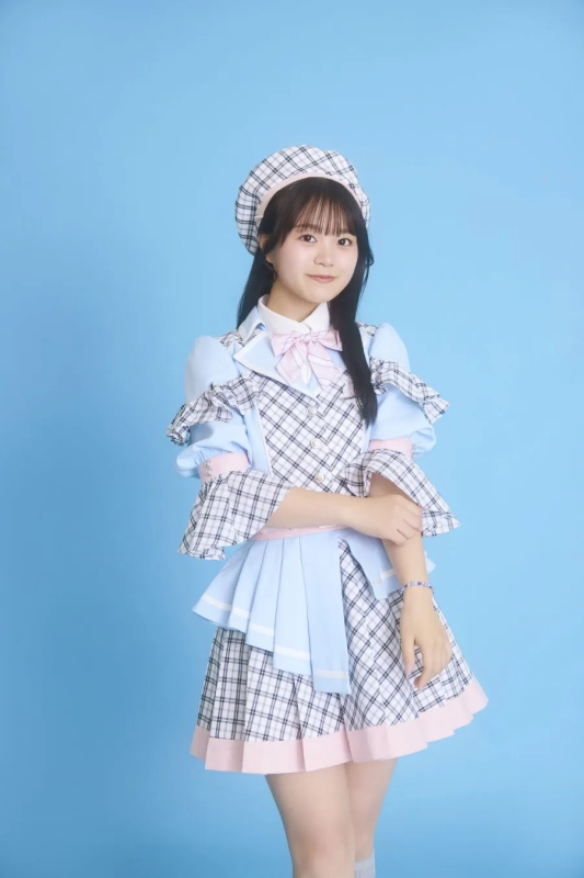 水色の背景に、チェック柄の衣装を着た若い女性が立っています。彼女はベレー帽をかぶり、腕を組んでカメラを見ています。