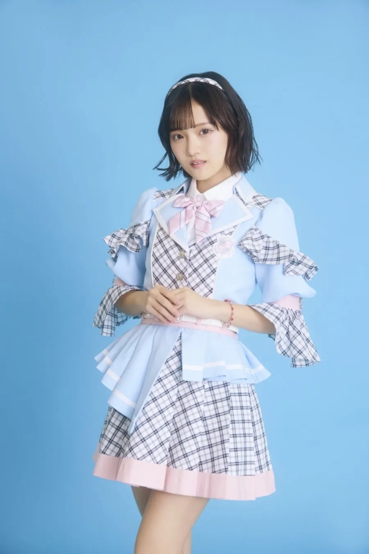 若い女性が、水色の衣装を着て、青い背景の前に立っています。彼女はカメラを見ています。