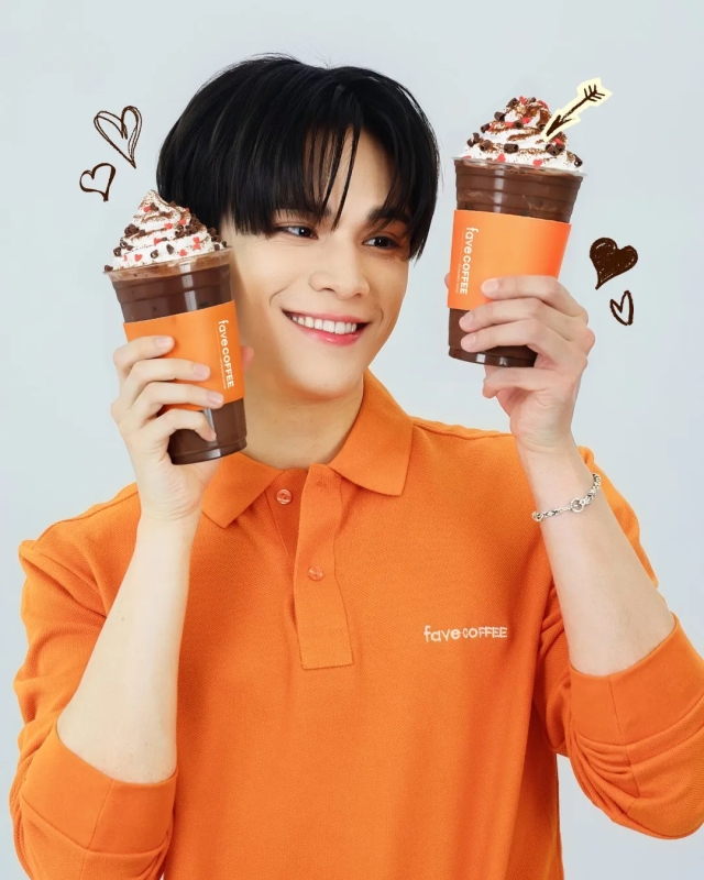 オレンジ色のポロシャツを着た男性が、2つのチョコレートドリンクを持ち、笑顔でポーズをとっている画像。ドリンクにはホイップクリームとトッピングが施されており、手書きのハートマークが描かれている。