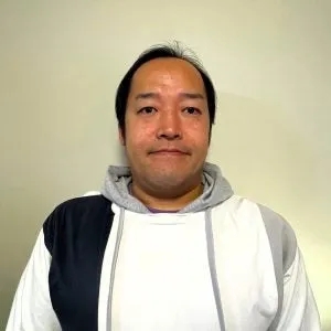 男性が正面を向いて立っている姿を捉えたポートレート写真です。男性は、白、グレー、紺色のフード付きのパーカーを着ています。背景は薄いベージュ色です。