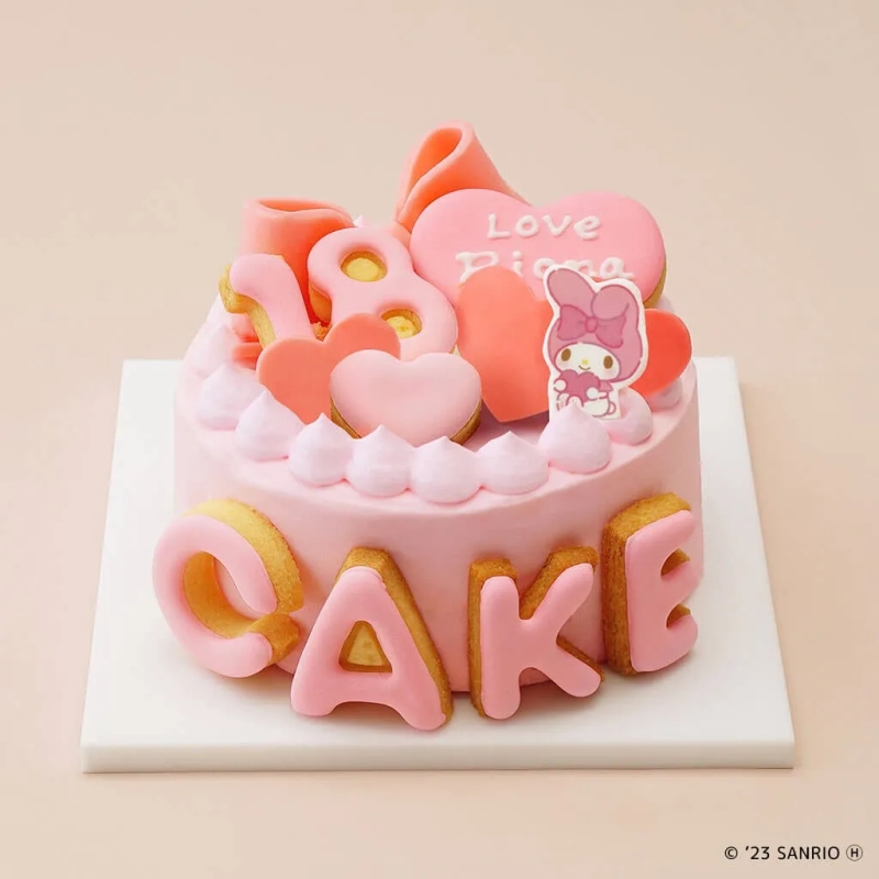 ピンク色のケーキ。上にはハートやキャラクターの飾り、側面にはCAKEの文字が施されている