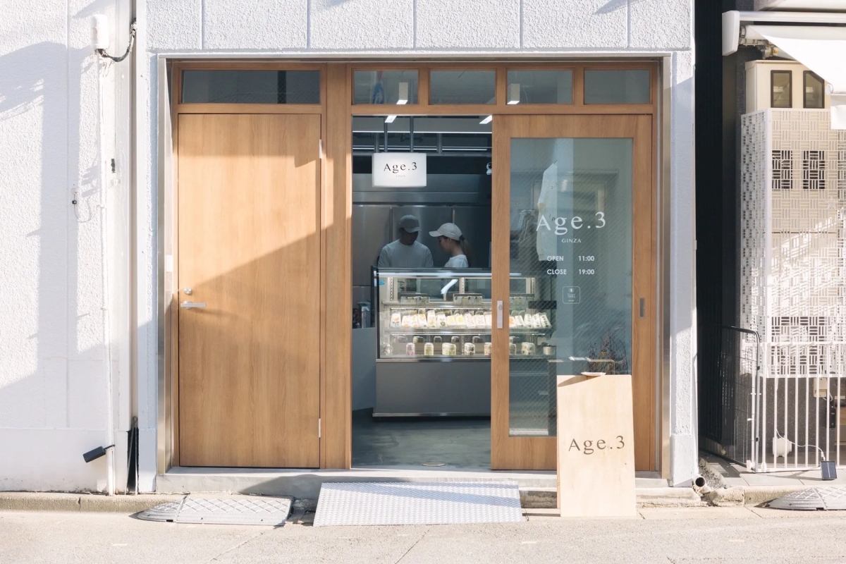 Age.3という店の外観の写真。木製のドアと窓があり、店員が中にいるのが見える。店の前には看板があり、営業時間が表示されている。