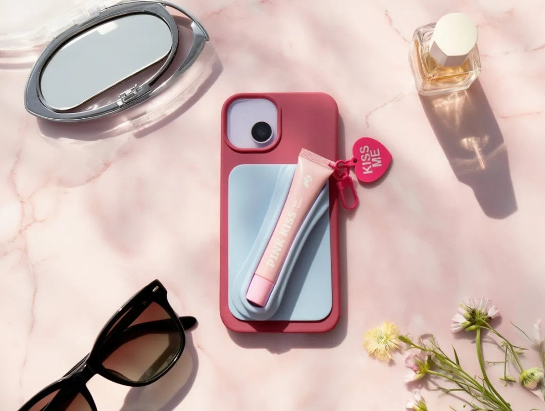 ピンク色の背景に、サングラス、化粧品、スマートフォンケース、鏡、香水が配置された写真です。