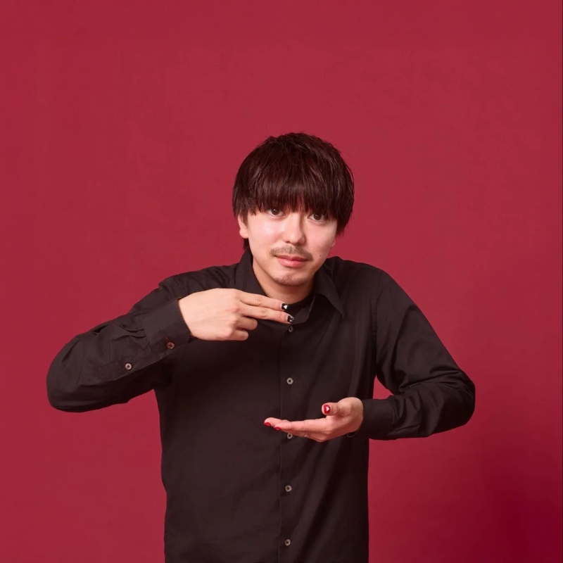 赤い背景の前で、黒いシャツを着た男性が、片方の手で何かを指し、もう片方の手を開いてそれを示している写真。彼の爪は黒と赤に塗られている。