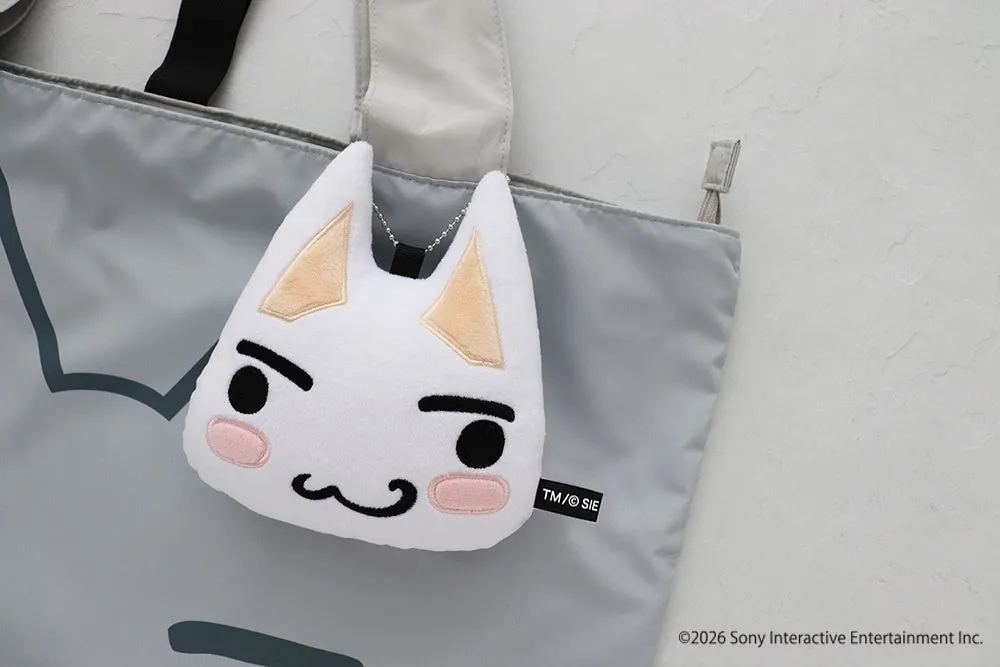 白い猫のぬいぐるみとグレーのバッグが写った写真。猫のぬいぐるみは、黒い目、眉毛、口、そして頬にピンク色のアクセントが施されており、バッグにぶら下がっている。バッグには黒い持ち手が付いており、背景は白い壁。