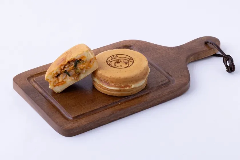 木製のまな板の上に、たい焼きのような丸い焼き菓子が2つ乗っている。一つは半分にカットされており、具材が見えている。もう一つは、キャラクターのイラストが描かれている。
