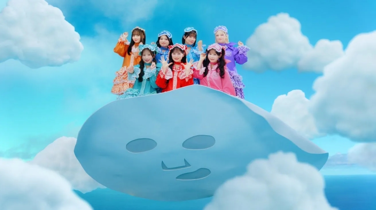 カラフルなフリル衣装をまとった7人の女性アイドルグループが、巨大なフェイシャルマスク型の乗り物に乗って青い空と白い雲の中にいる。皆が笑顔で手を振っており、夢のようなポップで可愛らしい世界観が広がる。