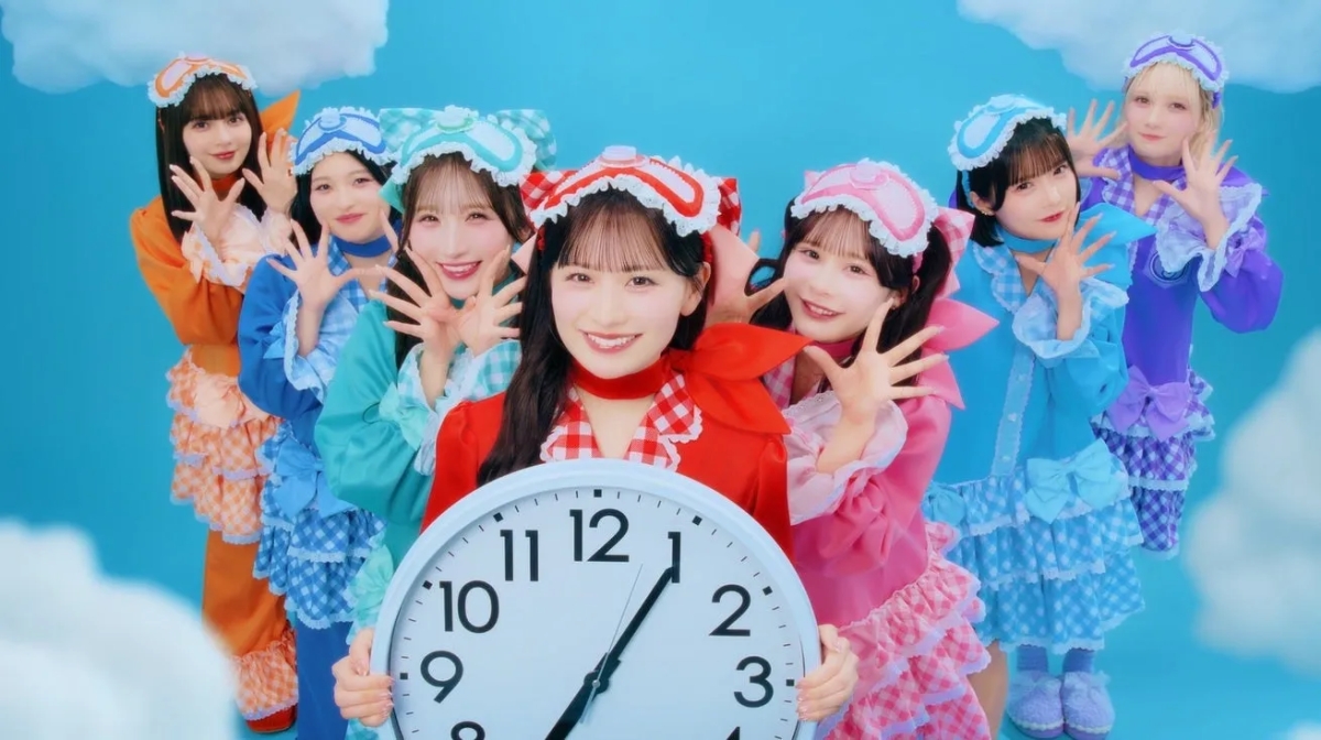 カラフルなパジャマ風衣装とスリープマスクを身につけた7人の若い女性アイドルグループが、笑顔でポーズを取りながら大きな時計を抱えています。背景は水色の空と白い雲で、全体的に可愛らしくポップな雰囲気の画像です。