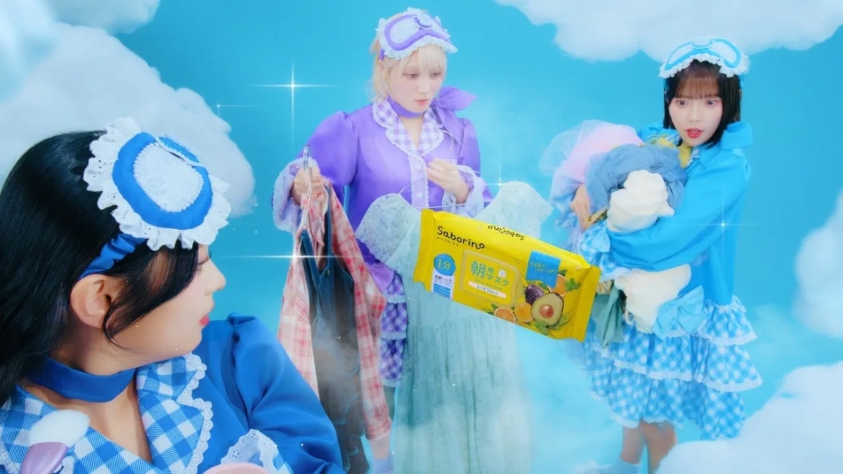 青い空と雲を背景に、パジャマとアイマスクを着用した3人の若い女性が写っています。中央の女性は黄色のサボリーノ朝用マスクを持っており、他の女性は服を抱えています。全体的に明るく、ファンタジーな雰囲気の商業写真です。
